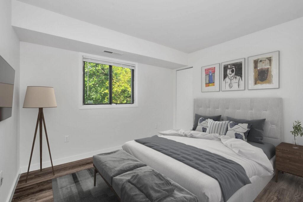 Experience the elegant charm of our luxury 2-bedroom apartments in Oxon Hill, featuring a minimalist design with a gray upholstered bed, white and gray bedding, serene views from the window, sleek wooden floors, abstract art adorning the walls, and a contemporary floor lamp. | Spark Living