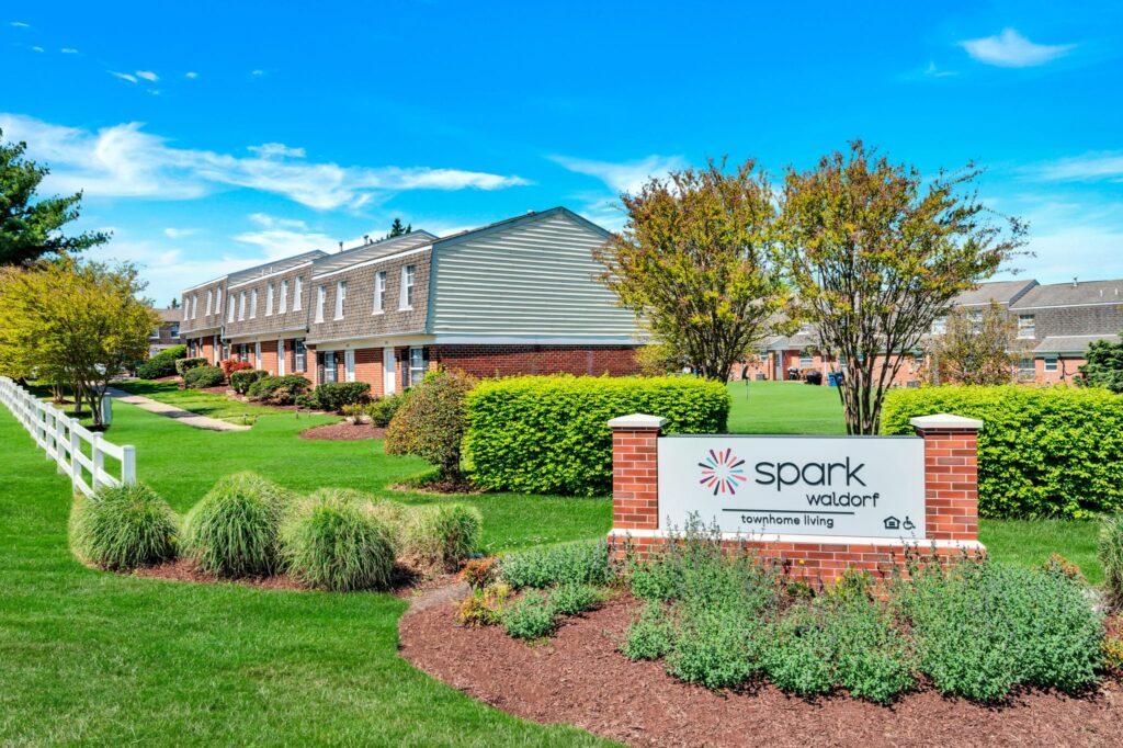 Brick townhouses with green lawns and trees under a blue sky in MD. A sign reads "Spark Waldorf, luxury 2-bedroom apartments and townhome living" in front. | Spark Living