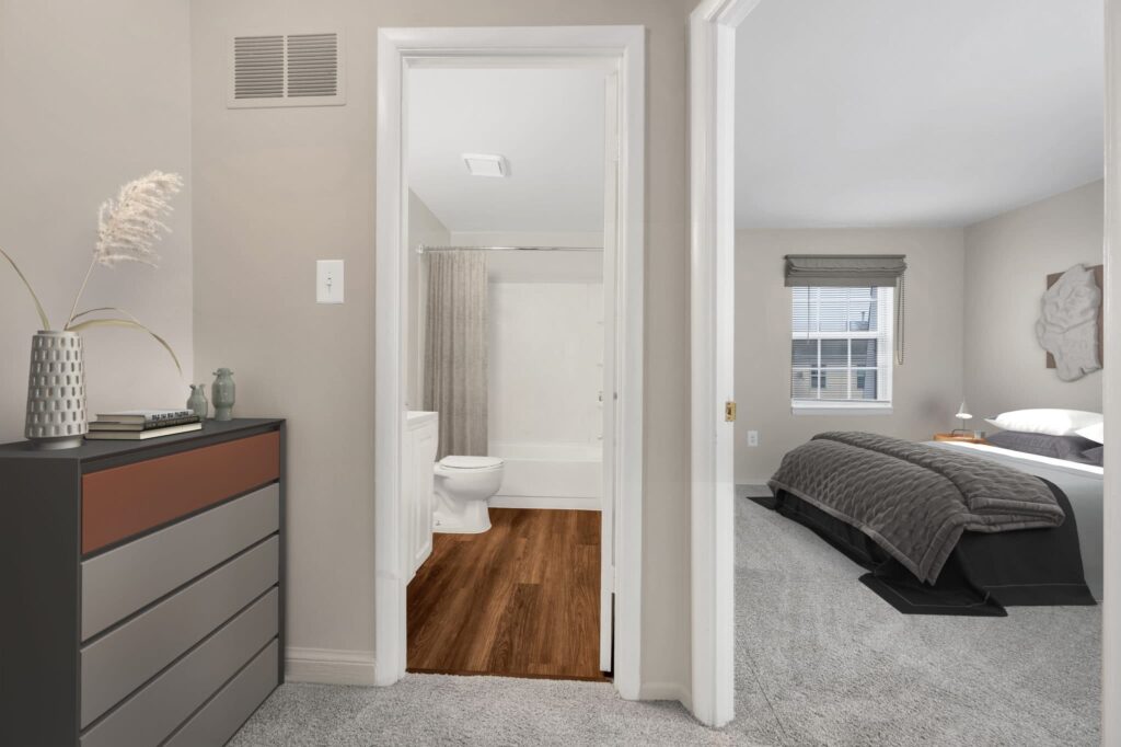 Experience the charm of luxury 2-bedroom apartments in Waldorf, MD. This elegant space features a bedroom with gray bedding and a stylish dresser adorned with books and a vase, seamlessly leading to a bathroom complete with a shower curtain and wooden floor. | Spark Living