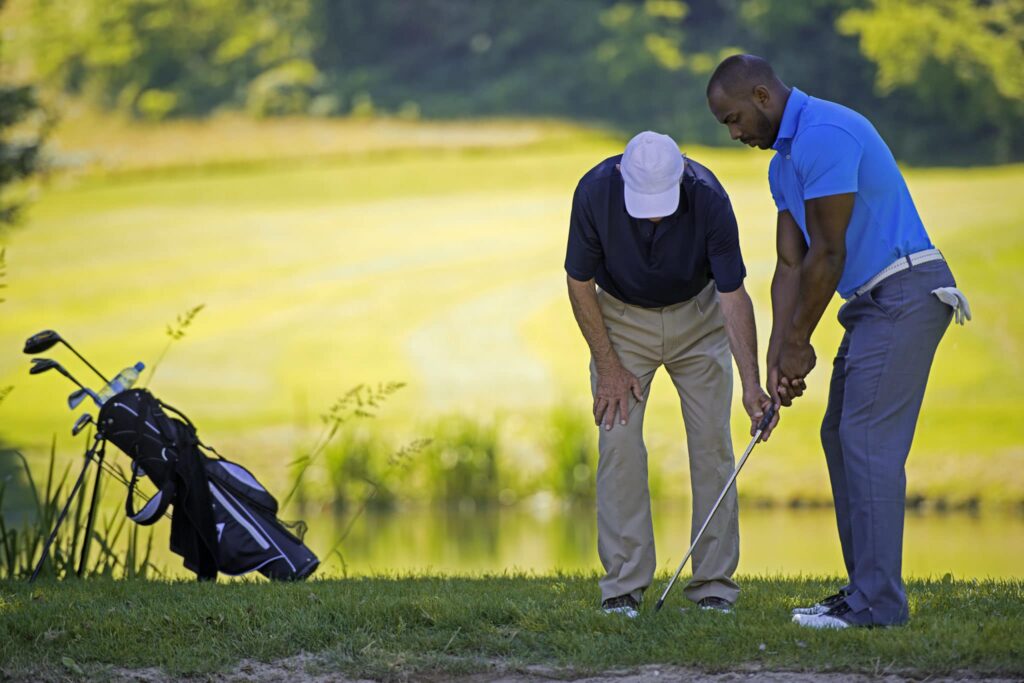 On a lush golf course, two men enjoy a lesson, one in blue guiding the other. A golf bag stands nearby, adding to the serene scene reminiscent of luxury living, much like the upscale 2-bedroom apartments in Oxon Hill, MD. | Spark Living